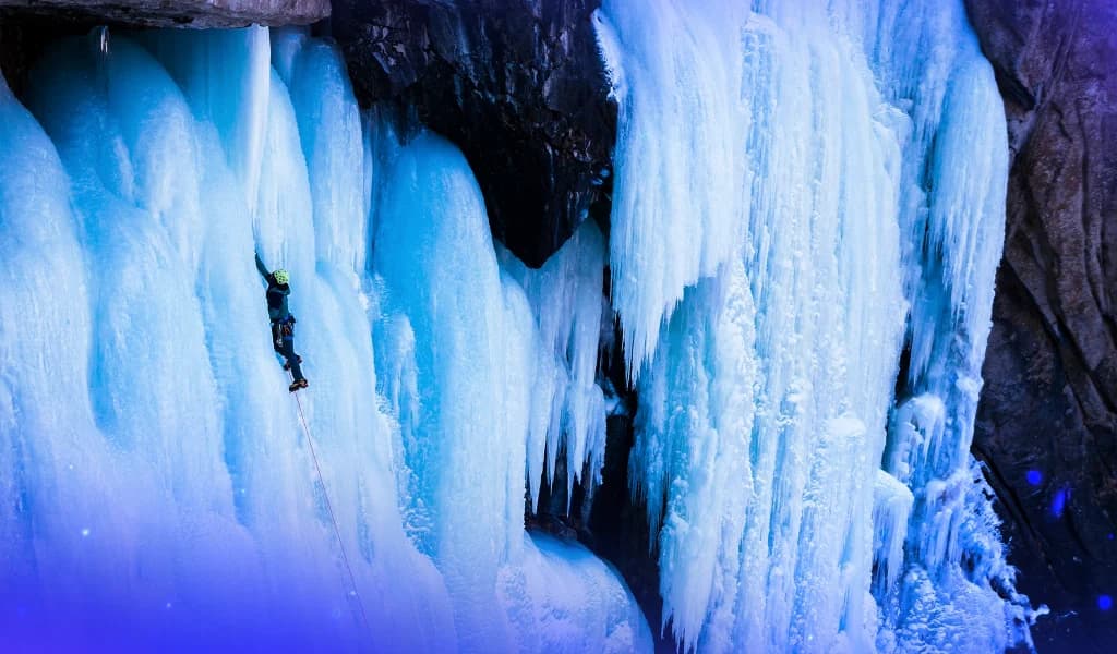 Climber wearing a helmet and harness scaling a tall frozen waterfall with ice axes.