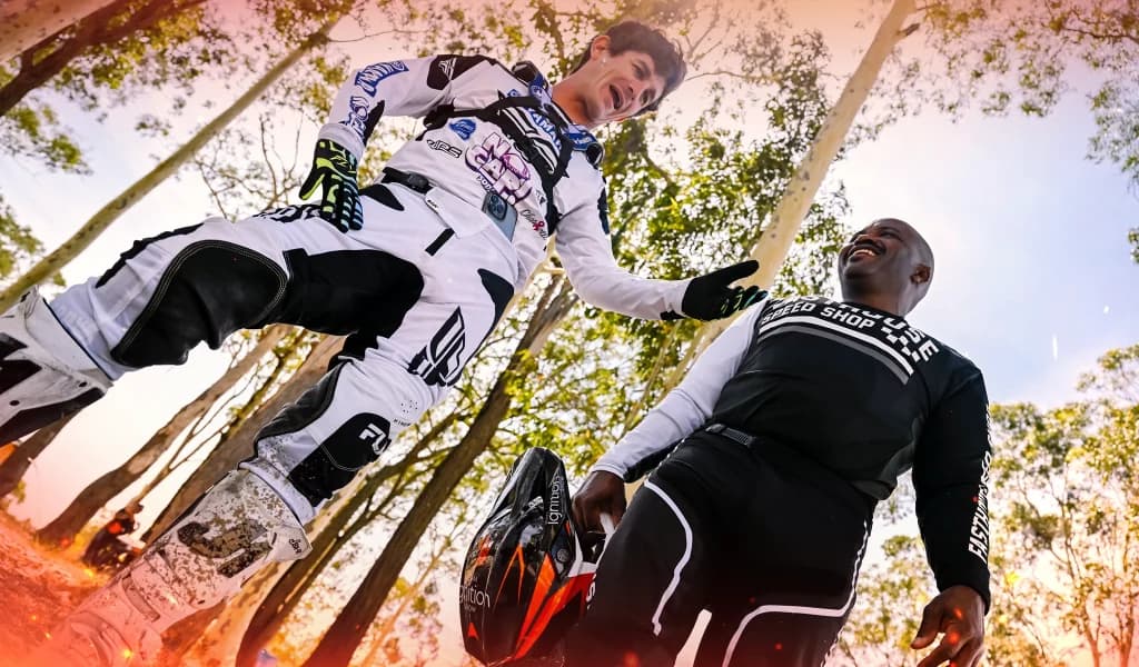 Two men, Jarryd McNeil and Selema Masekela, in motocross gear stand outdoors smiling and talking, one holding a helmet near a dirt track.