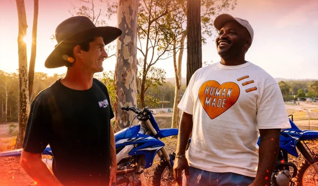 Two men, Jarryd McNeil and Selema Masekela, seated outdoors during a recorded conversation, with microphones set up between them near a dirt track.