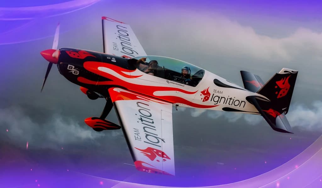 Red, white, and black aerobic airplane wrapped in Team Ignition branding flying at a steep angle through a cloudy sky.