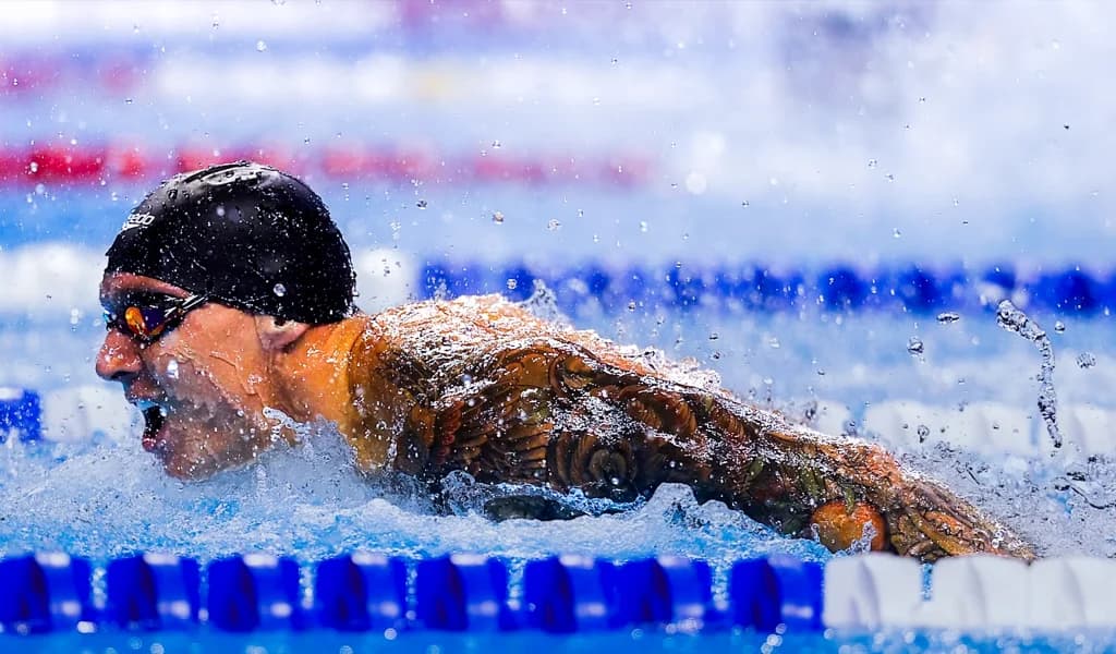 Caeleb Dressel swimming mid-race, goggles and cap on, water splashing around him.