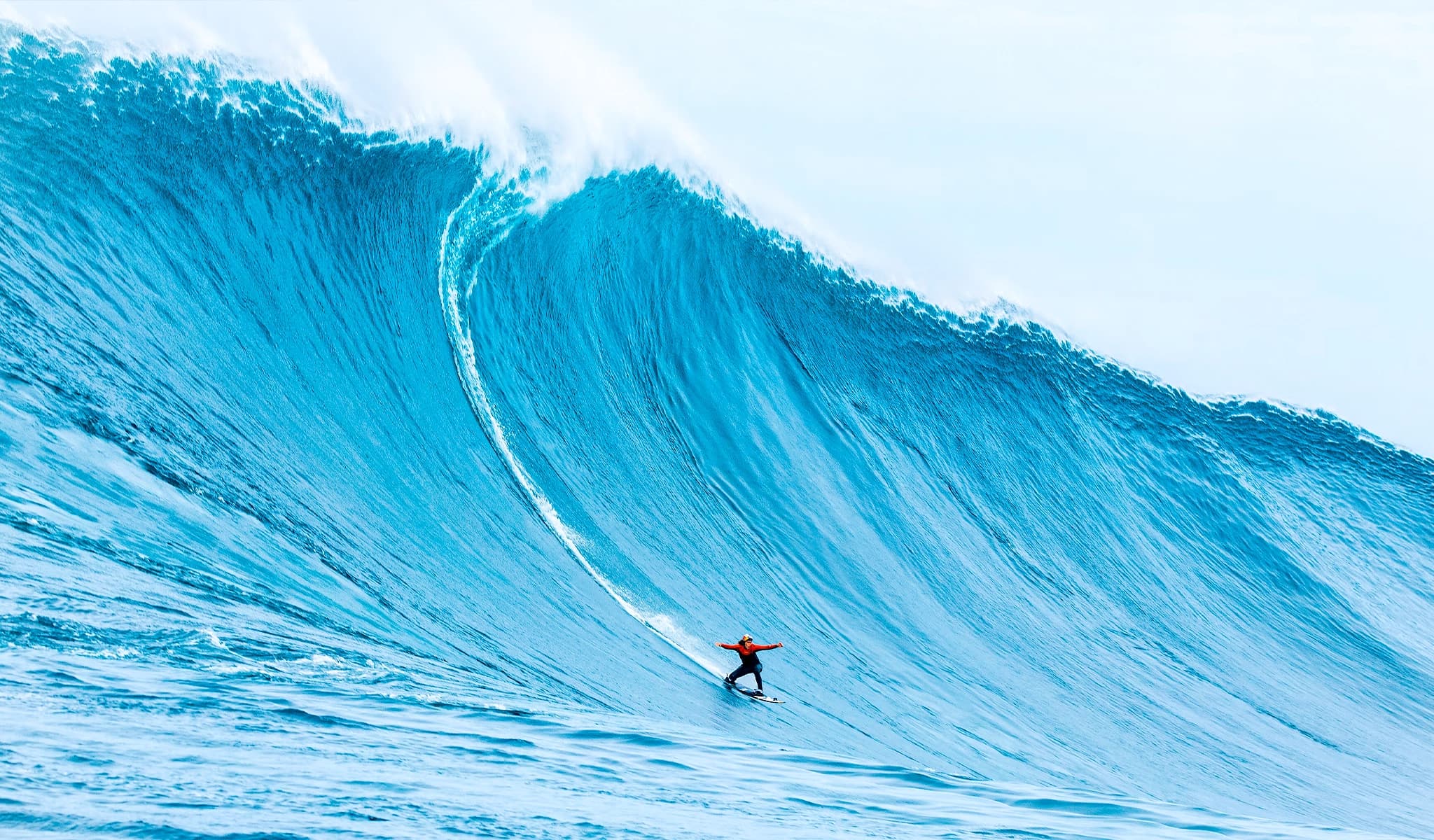 A surfer rides a massive blue wave with arms outstretched, dwarfed by the wall of water.