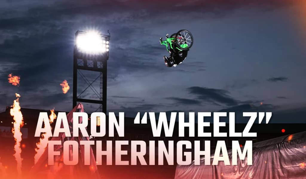 WCMX athlete Aaron Wheelz Fotheringham in a wheelchair completing a backflip under stadium lights with flames in the background.