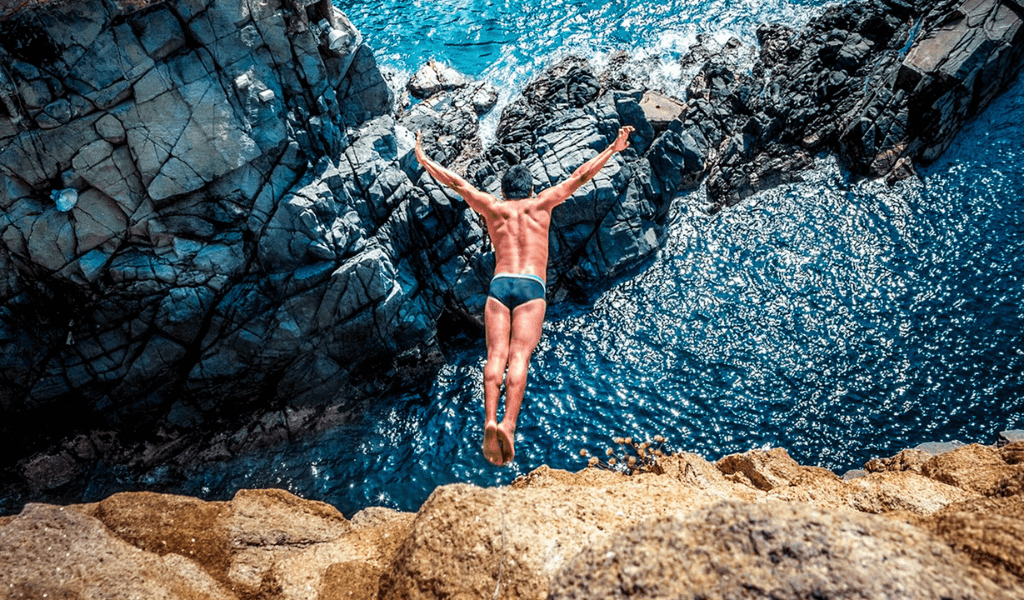 Overhead view of a male diver leaping arms wide from a rock ledge toward the ocean below.