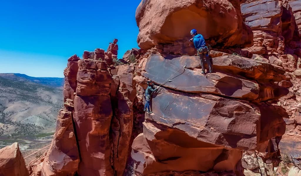 JJ Yosh and another climber on a red rock face connected by a rope with a canyon valley visible in the background.