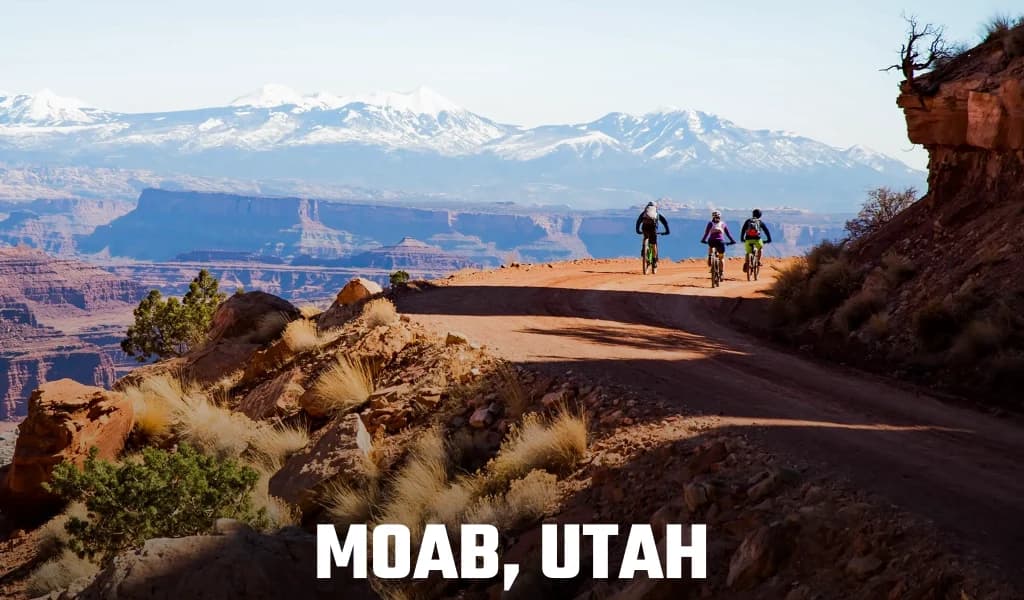 Text reading Moab, Utah over three mountain bikers riding a cliffside trail with snow-capped mountains behind them.