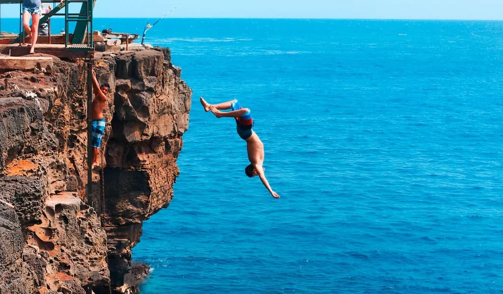 A cliff jumper flips head-first off a rocky ledge into deep blue ocean water.