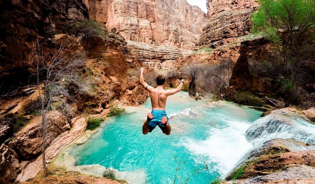 A jumper leaps feet-first with arms raised into a turquoise canyon pool below.