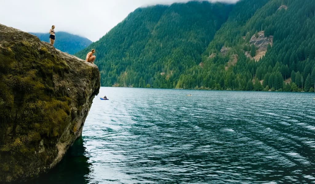 Two people stand atop a mossy cliff over a mountain lake, one crouched to check the drop.