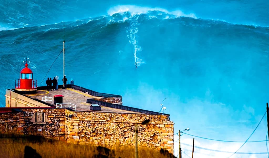 A massive wave breaks behind the red Nazaré lighthouse as spectators watch from the fort.