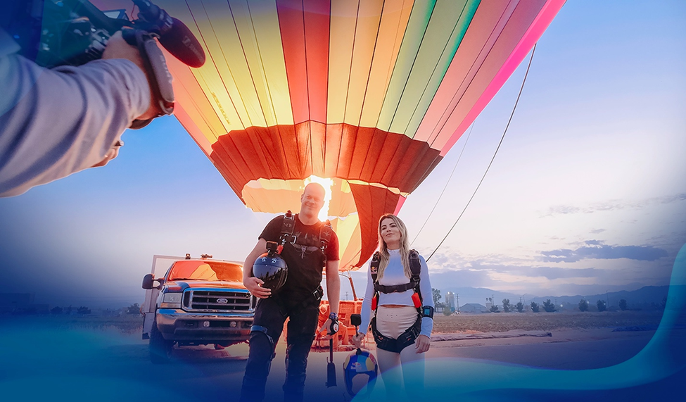 Jeb Corliss and Leticia Bufoni standing in front of a colorful hot air balloon, preparing for a skydive, with The Team Ignition Show podcast camera crew capturing the moment.