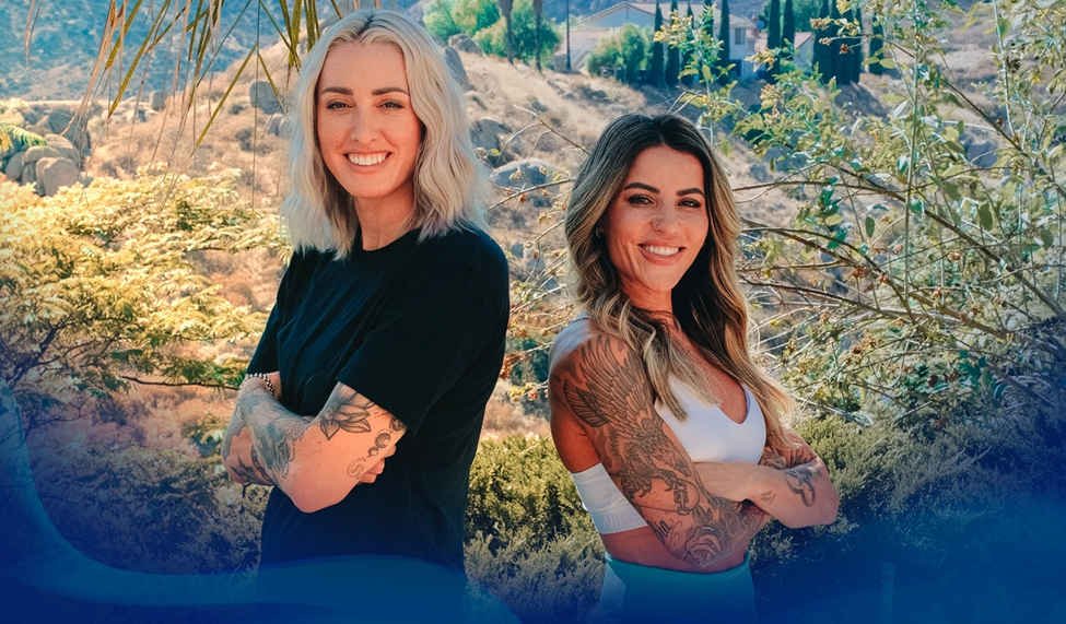 Alex Johnson and Leticia stand back-to-back outdoors, smiling confidently at the camera, with trees and a mountain in the background, showcasing their arm tattoos.