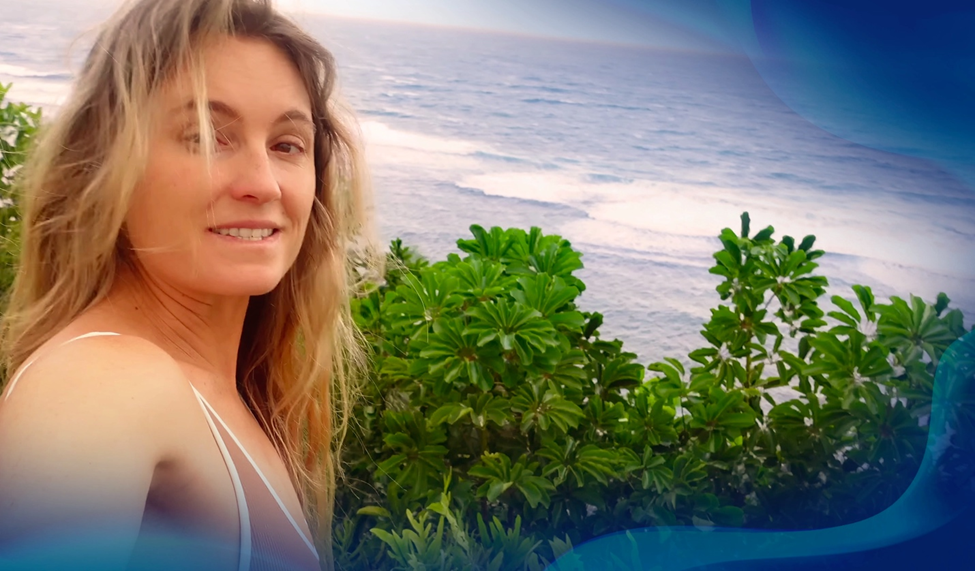 Alana Blanchard smiling and taking a selfie near the coastline, with green plants and the ocean in the background.