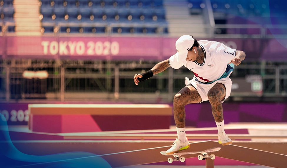 A skateboarder performing a trick in the 2020 Tokyo Olympics, mid-air with blurred stadium seating in the background.