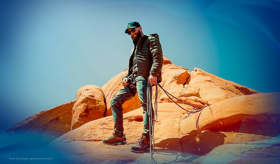 Ryan Robinson in outdoor gear, holds ropes while standing on a rock formation under a clear blue sky, preparing for a highline setup.