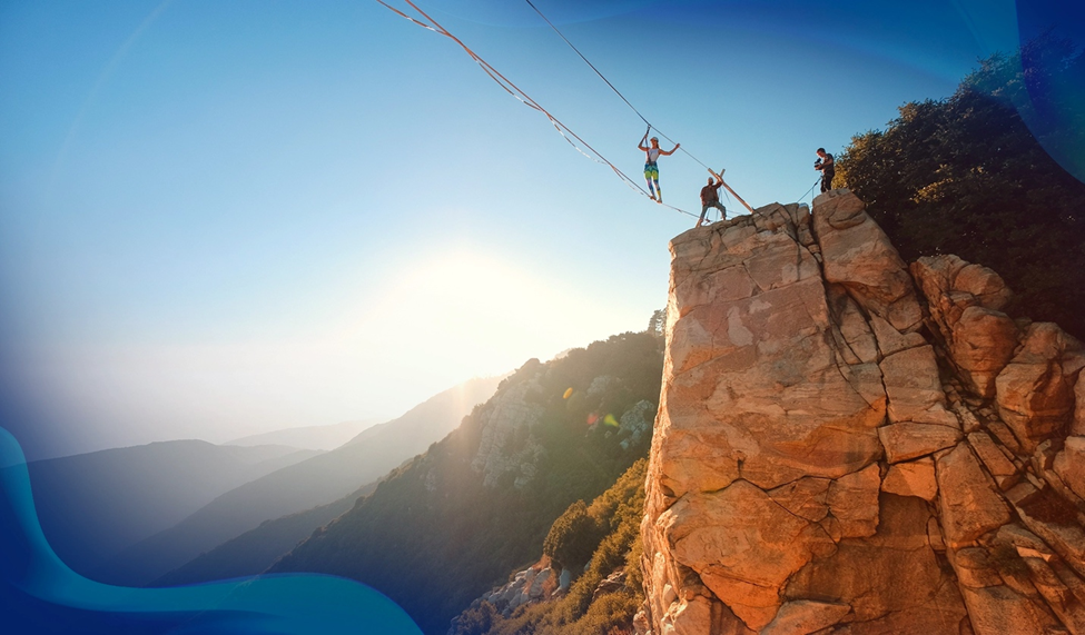 A highliner balances on a slackline stretched between two cliffs, with a golden sunset illuminating the rocky landscape and distant hills.