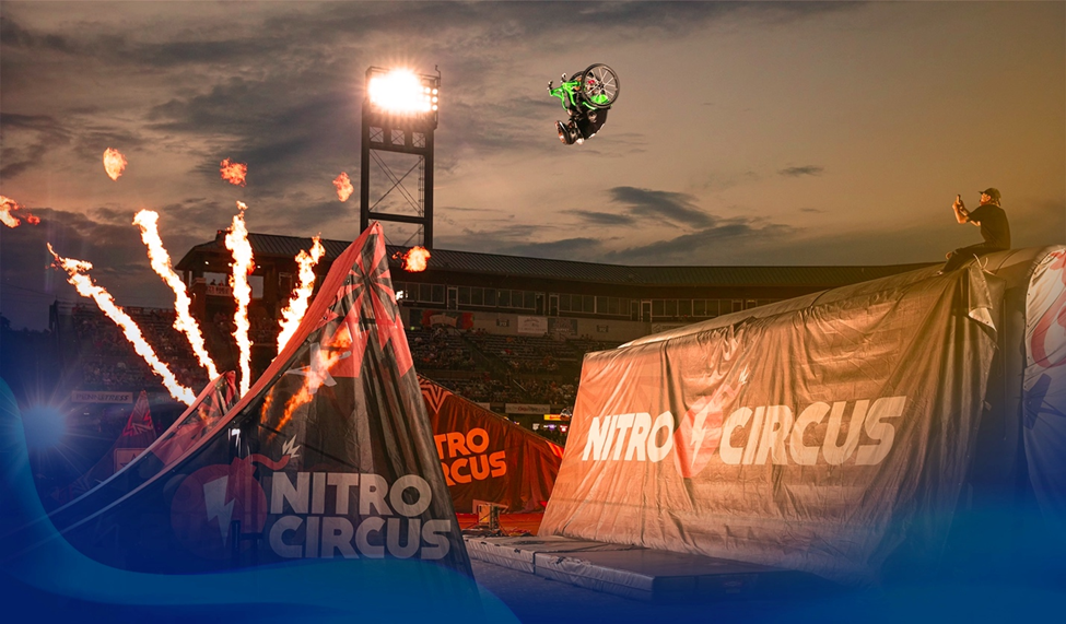 Wheelchair motocross athlete mid-air performing a backflip against a backdrop of fiery ramps and a sunset-lit Nitro Circus stadium.