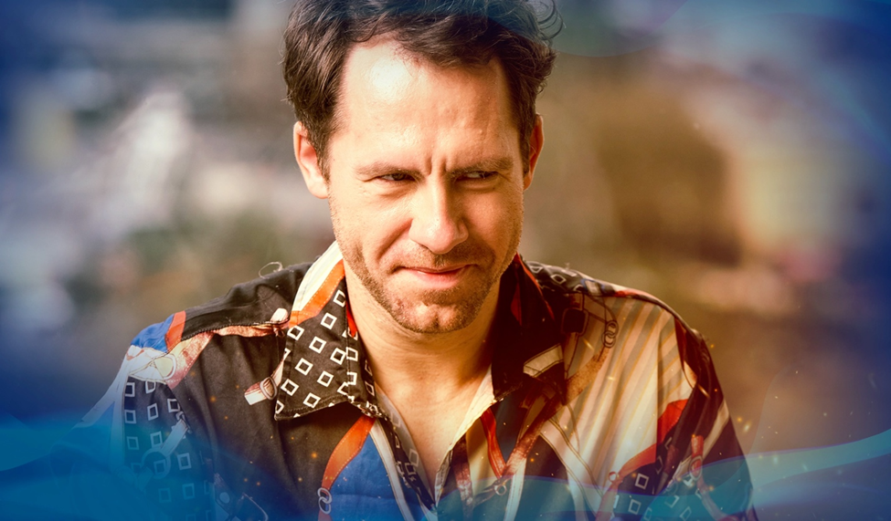 Daniel Cates, wearing a colorful, patterned shirt, smirks confidently, sitting in a well-lit room with a blurred background