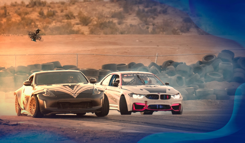 Two race cars drifting on a track surrounded by tires, creating a smoky trail under a warm sunset sky.