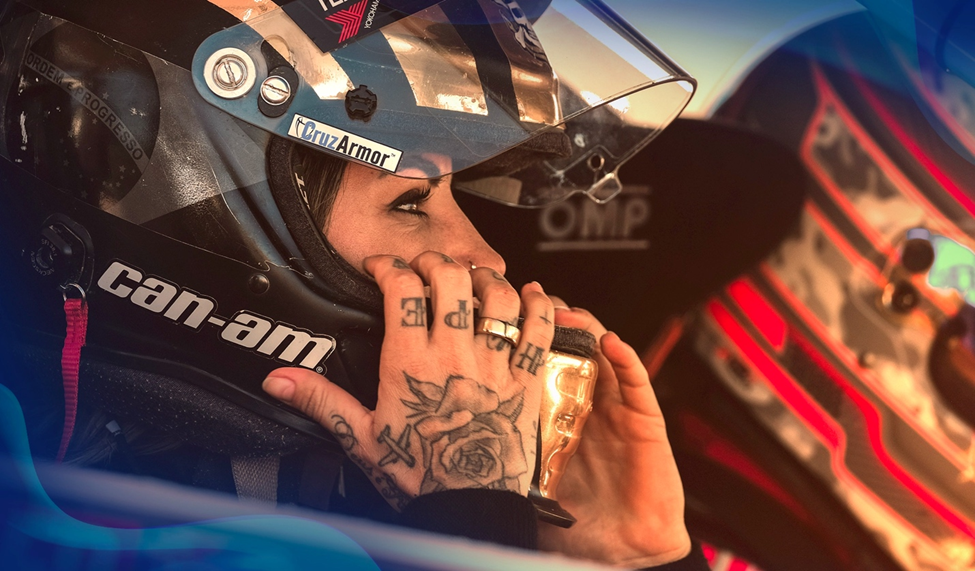 Leticia Bufoni gripping her helmet inside a race car, tattoos on her hand visible, with a calm yet focused expression.