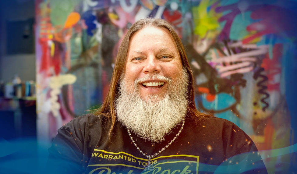 Graffiti artist RISK smiling in front of a colorful graffiti wall, wearing a black shirt with a white beard.