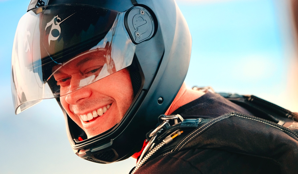 Close-up of Jeb Corliss wearing a helmet, smiling, with a blurred blue sky background. Frame from his appearance in the Team Ignition Show.